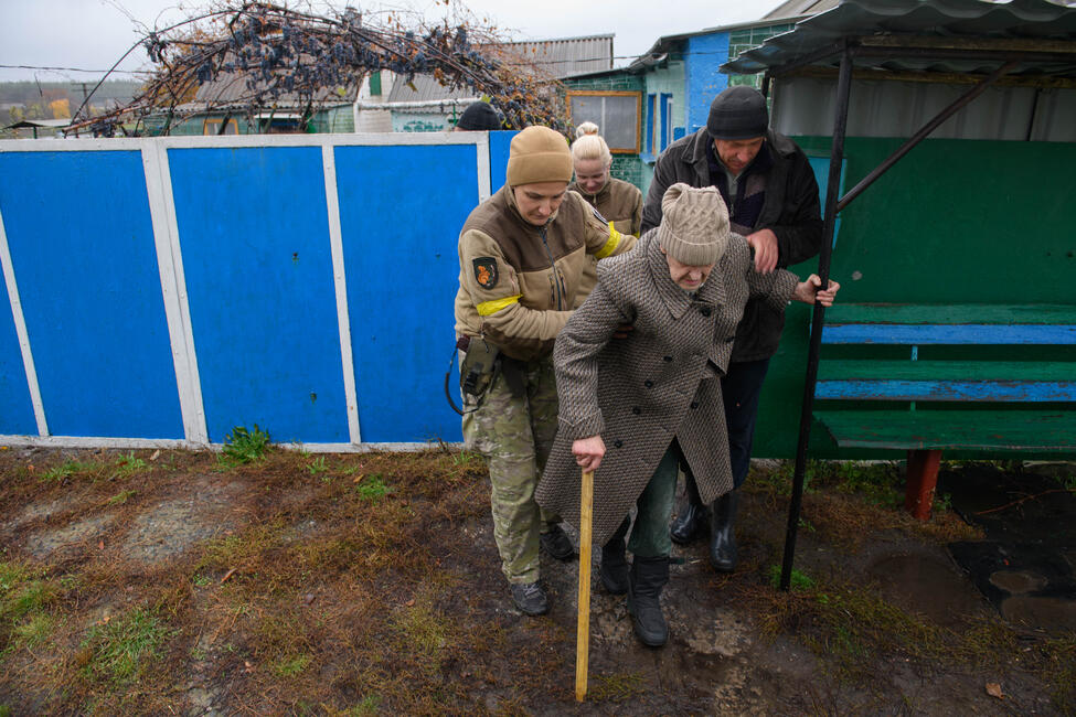 Eine ältere Frau mit Gehstock wird beim Verlassen ihres Grundstücks von einer Soldaten in Uniform und einem Mann in Zivilkleidung gestützt. Alle abgebildeten Personen tragen Winterkleidung.