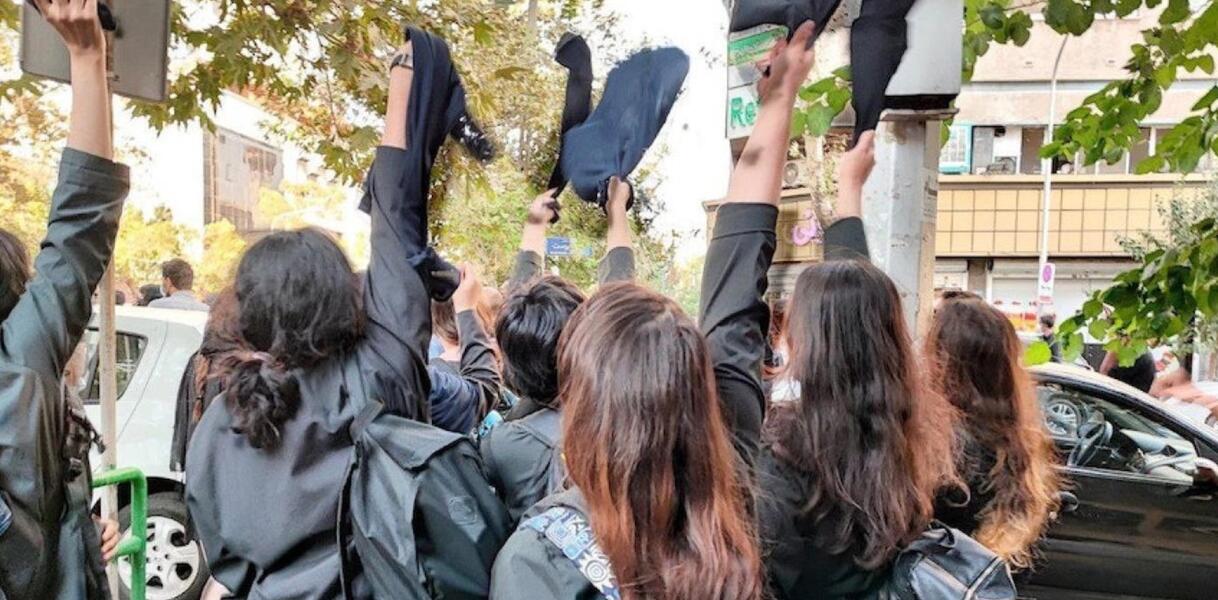 Das Bild zeigt mehrere junge Frauen von hinten, die schwarze Kopftücher in die Luft werfen