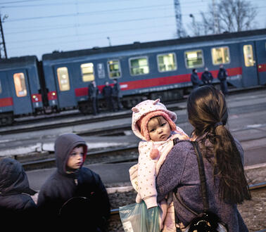 Eine Mutter mit Kindern steht auf einem Gleis am Bahnhof, im Hintergrund hält ein Zug, vor dem Menschen stehen.