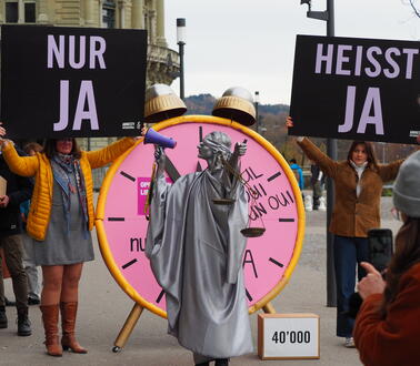 Das Bild zeigt eine Justitia-Figur, die ein Megaphon in der Hand hält. Hinter hier stehen Menschen, die große Plakate hoch halten auf denen "Nur Ja heißt Ja" steht. 