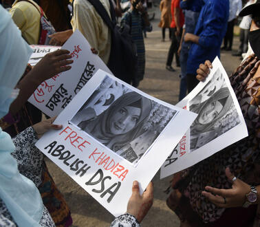 Eine Frau steht in einer Menschenmenge. Sie hält ein Plakat in ihren Händen und betrachtet ist. Auf dem Plakat ist ein Porträtfoto von Khadijatul abgedruckt, auf dem sie ein Kopftuch trägt. Unter dem Foto steht unter anderem: "Free Khadiza".