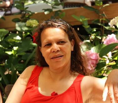 Eine Frau mittleren Alters sitzt bei Sonnenschein draußen for Rosensträuchern, sie trägt ein schulterloses Top.