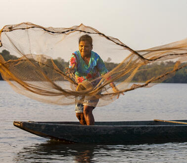 Das Bild zeigt eine Fischerin auf einem Boot, die gerade ein Netz auswirft
