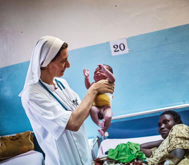 Eine Krankenschwester hält ein Neugeborenes in den Händen, im Hintergrund die Mutter des Kindes in einem Krankenhausbett, an der Wand über dem Bett hängt die Nummer 20.