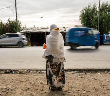 Das Foto zeigt eine Frau am Rande einer Straße, auf der Autos fahren. Sie trägt ein Gewand und ein Kopftuch und trägt ein Kind auf ihrem Rücken.