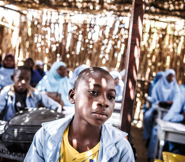 Ein Junge sitzt in einer Schule in Niger, einem Gebäude aus Holzzweigen, durch dessen Wände Sonne fällt, hinter ihm sitzen andere Schüler*innen, die Mädchen tragen Kopftuch.