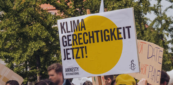 Das Foto zeigt eine dichtgedrängte Menschenmenge auf einer Demonstration. Eine junge Frau mit Amnesty-Mütze hält ein Schild mit Amnesty-Logo hoch. Auf dem Foto steht: "Klimagerechtigkeit jetzt!".