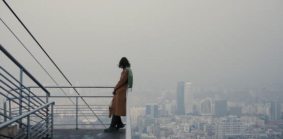 Eine junge Frau im Mantel steht auf einer Plattform im Freien, im Hintergrund eine Stadt.