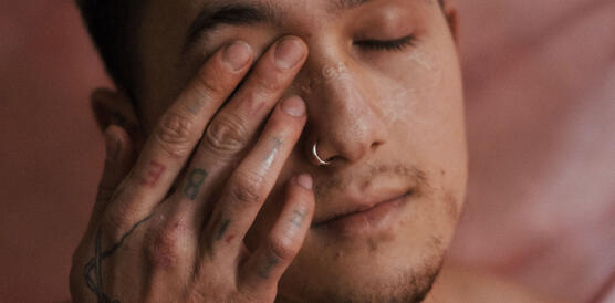 Ein junger Mann, kurzes blaugefärbtes Haar, die Augen geschlossen, Oberkörper nackt, Tatoos auf dem Körper, er reibt sich mit der rechtenHand das rechte Auge.