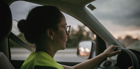Eine junge Frau mit Brille, die Haare zurückgebunden, trägt eine neongelbe Warnweste, sitzt am Steuer eines VW-Autos und fährt.