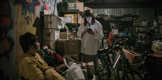 Eine stehende junge Frau am Handy in einem vollgestellten Lagerraum, Fahrräder, Kisten, Gerümpel, auf einem Sofa sitzt ein junger Mann.