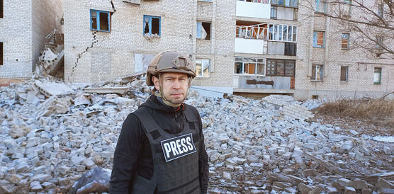 Ein Mann mit Soldatenhelm und Schussweste, auf der "Press" steht, befindet sich vor einem durch Raketenbeschuss zerstörten Gebäude.