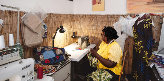 Eine nigerianische Frau arbeitet in einer Schneiderwerkstatt an einer Nähmaschine, eine kleine Lampe brennt über ihrem Arbeitsplatz, um sie herum hängen Stoffe und Kleider.