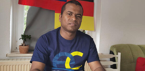 Ein Mann sitzt an einem Schreibtisch vor einem Dokument, hinter ihm hängen zwei Flaggen, die venezolanische und die deutsche.