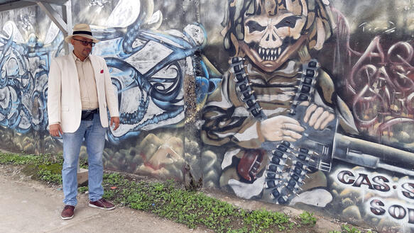 Ein Mann mit weißem Jacket und weißem Hut steht vor einer Mauer, die mit Grafitti bemalt ist.