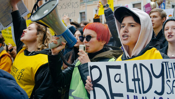 Verschiedene Menschen, darunter Amnesty-Unterstützende mit gelben Westen und dem Amnesty-Logo demonstrieren, eine Frau trägt ein Megaphon. 