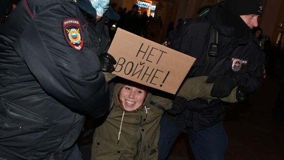Eine Frau hält ein Pappschild mit russischer Schrift in der Hand, während zwei Polizisten sie an den Armen packen und wegzerren.