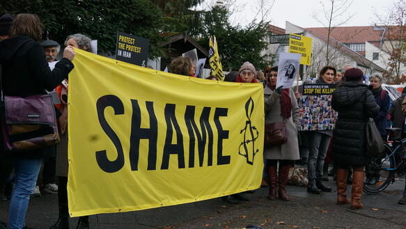 Das Bild zeigt mehrere Menschen mit einem Plakat, auf dem steht "Shame"