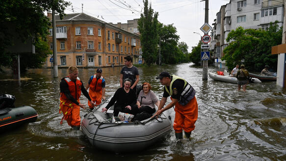 Das Bild zeigt ein Schlauchboot, überflutete Straßen, zwei ältere Menschen befinden sich im Schlauchboot