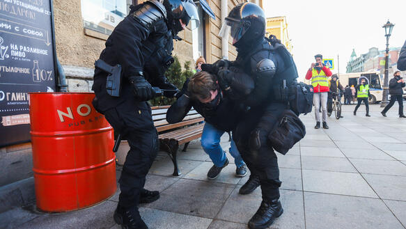 Das Bild zeigt zwei schwer bewaffnete Polizisten, wie sie eine Person festhalten und den Kopf zu Boden drücken.