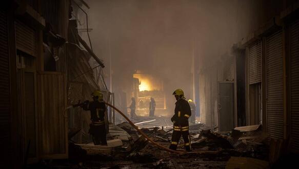 Die Nachtaufnahme zeigt Feuerwehrleute in einer nur durch Feuer erleuchteten Straße mit zerstörten Gebäuden