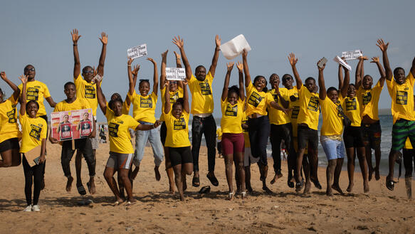 Das Bild zeigt viele junge Menschen mit gelben T-Shirts an einem Strand, sie reißen die Arme in die Luft, viele lachen