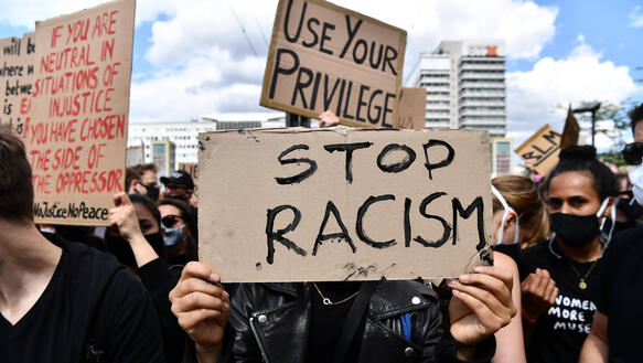 Das Bild zeigt viele Menschen mit Protestplakaten, auf einem steht "Stop Racism"