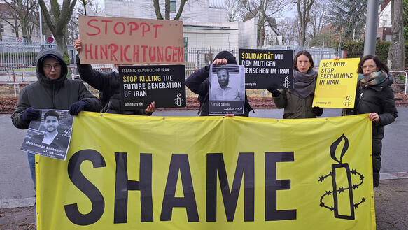 Das Bild zeigt mehrere Menschen mit Protestplakaten, eines mit dem Schriftzug "Shame"