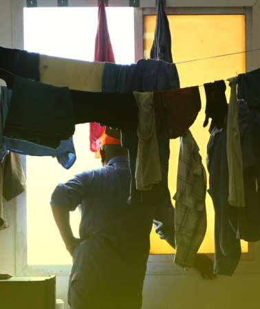 In einem dunklen Raum, in dem Wäscheleinen mit Wäsche hängen, schaut ein Mann mit dem Rücken zum Fotografen stehend aus dem Fenster. Das Foto wurde in einer Unterkunft für Arbeitsmigrant*innen in Katar aufgenommen, die dort für die WM Katar 2022 arbeiten.
