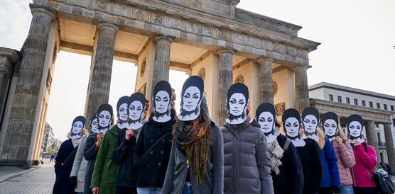 Vor dem Brandenburger Tor stehen 12 Frauen und bilden eine Pfeilformation, sie tragen Pappmasken mit dem Gesicht von Eren Keskin