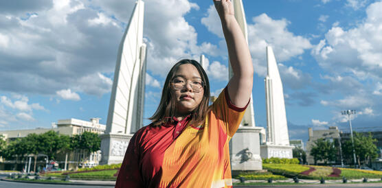 Eine junge thailändische Aktivistin steht vor einem Denkmal und hebt ihre linke Hand in den Himmel, über den Wolken treiben.