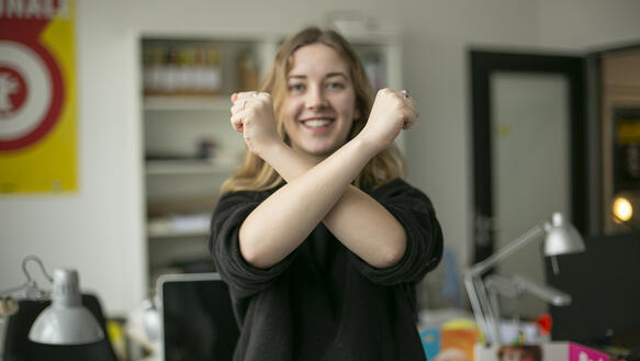 Eine junge Frau steht in einem Büro, lächelt in die Kamera und formt mit ihren Unterarmen ein X.