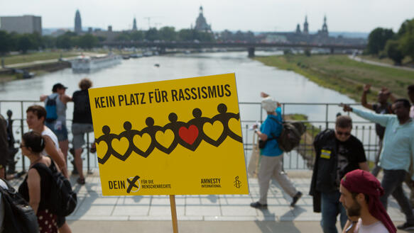 Ein Demoschild auf dem Steht "Kein Platz für Rassismus". Im Hintergrund ist ein Fluss zu sehen und es laufen ein paar Menschen über eine Brücke. 