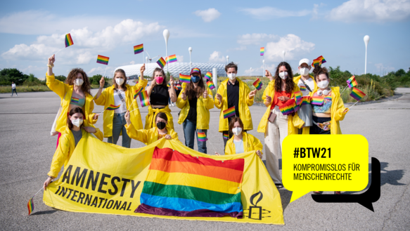 Eine Menschengruppe in gelben Jacken wedeln mit Regenbogenflaggen, sie haben vor sich ein gelbes Transparent auf dem ebenfalls die Regenbogenflagge ist und auf dem "Amnesty International" steht. Im Hintergrund die Allianz-Arena in München. Rechts unten in der Ecke eine gelbe Sprechblase, darin steht in schwarzer Schrift: #BTW21. Komromisslos für Menschenrechte