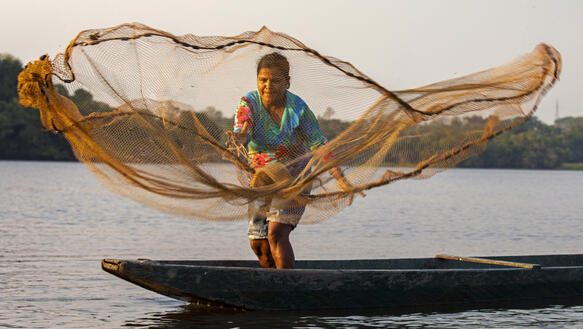 Das Bild zeigt eine Fischerin auf einem Boot, die gerade ein Netz auswirft