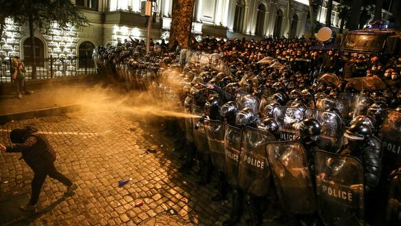 Das Foto zeigt dutzende Polizeikräfte mit Helmen und Schilden, die eng aneinander stehen. Aus der vorderen Reihe wird Tränengas auf einen Demonstranten gesprüht, der wegläuft. Das Foto entstand nachts.