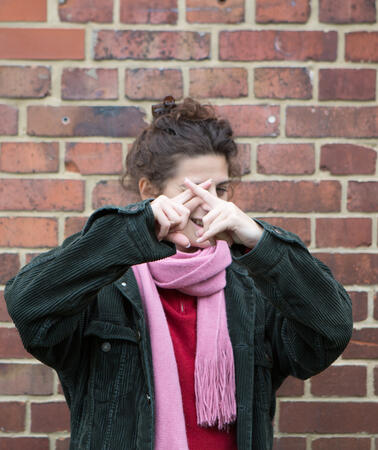 Eine junge Frau steht vor einer roten Backsteinwand, sie hält ihre Hände vor ihr Gesicht und formt mit ihren Zeigefingern ein Kreuz