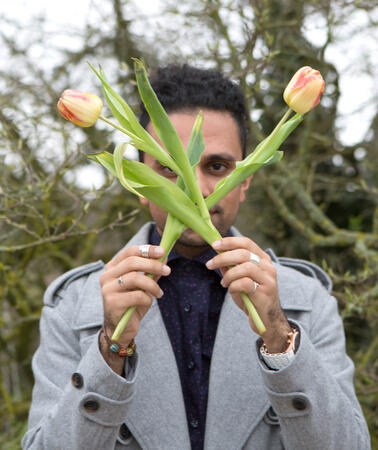 Ein junger Mann steht vor einem Baum und blickt in die Kamera. In seinen Händen hält er zwei rot-gelbe Tulpen, mit denen er vor seinem Gesicht ein Kreuz formt.