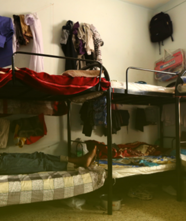 Männer liegen auf drei Stockbetten aus Metall. Diverse Kleidungsstücke hängen an den Wänden und an den Betten.