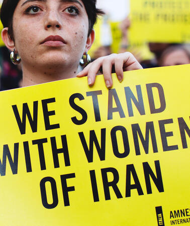 Das Bild zeigt eine Frau, die ein Schild in der Hand hält: "We stand with Women of Iran"